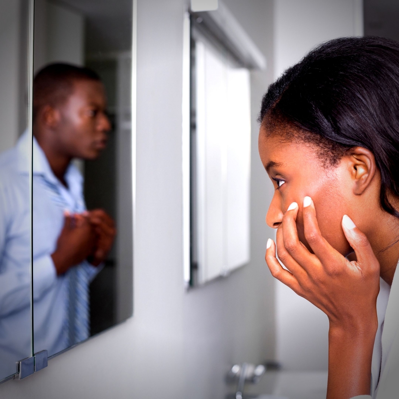 Foto de uma mulher negra, com uma das mãos no rosto, que olha para um espelho que está na sua frente. Ele reflete a imagem de um homem sério, que usa uma blusa social e está com as mãos na blusa, perto da gola.