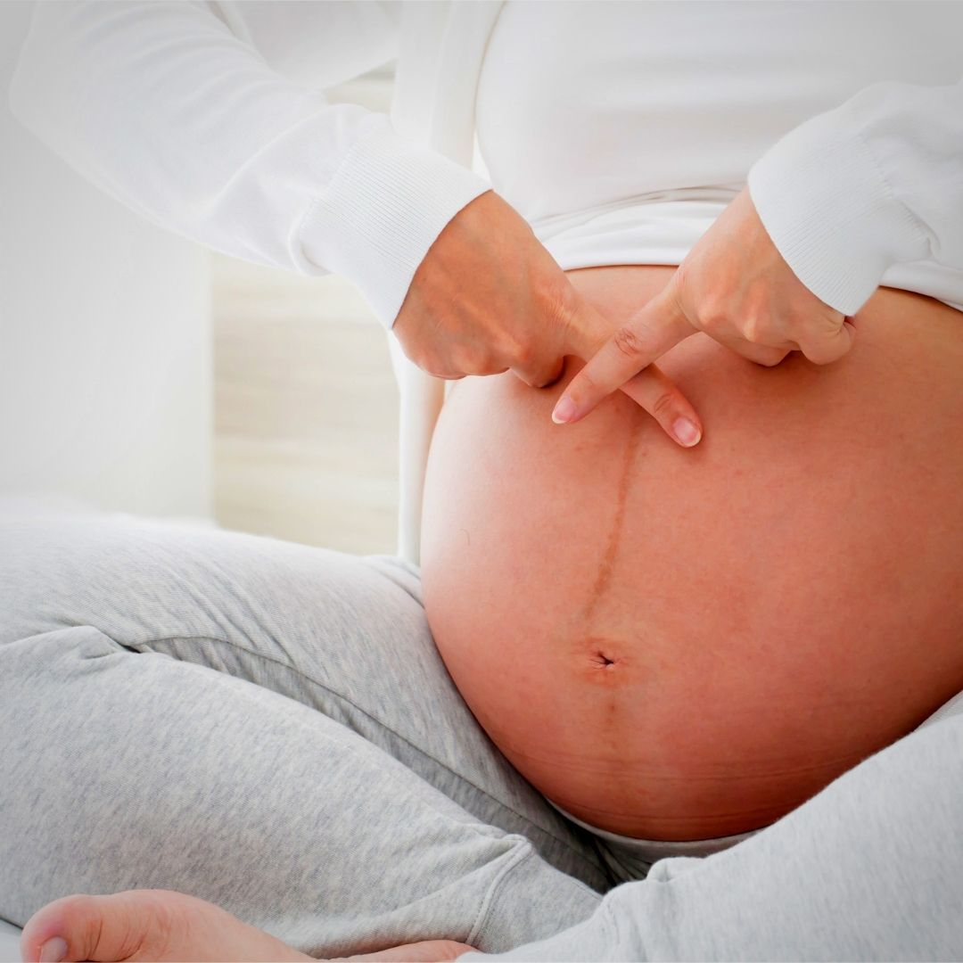 Foto de mulher grávida sentada, com a barriga de fora e os dedos indicadores cruzados em cima dela em forma de X.